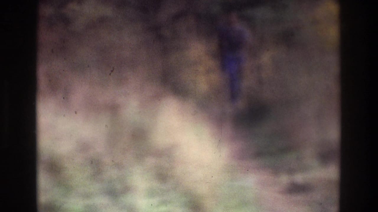 Soft-focused 1975 Atlanta 8mm frame showing a distant man in blue moving through tall grass in an open field or park-like area.