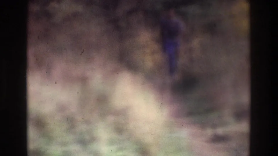 Soft-focused 1975 Atlanta 8mm frame showing a distant man in blue moving through tall grass in an open field or park-like area.