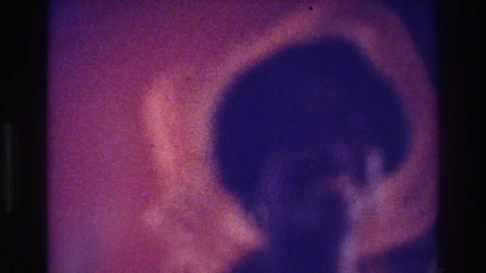 1975 Bronx home-movie frame with a close, blurred straw hat and adult figure, marked by pink-purple aging and heavy grain.