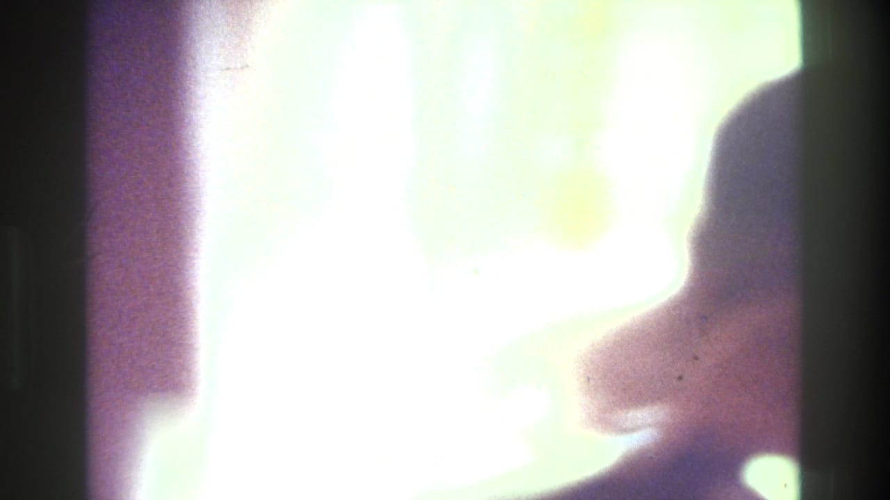 1975 Bronx home-movie frame showing a softly backlit woman in profile near a mirror, with bright window light and dark interior edges.