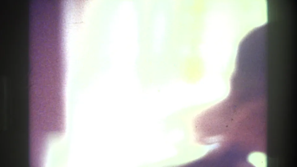 1975 Bronx home-movie frame showing a softly backlit woman in profile near a mirror, with bright window light and dark interior edges.