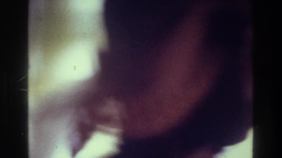 Soft 8mm frame from a 1975 Bronx home interior, showing a blurred close view of a woman in pink-toned clothing.
