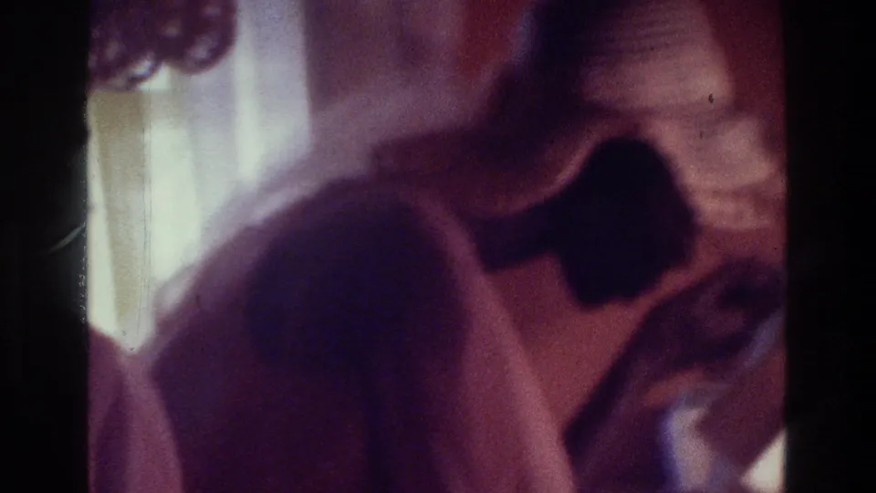 1975 Bronx home-movie frame showing a woman in a pink dress near a mirror, with a dark square detail on the garment and soft interior light.