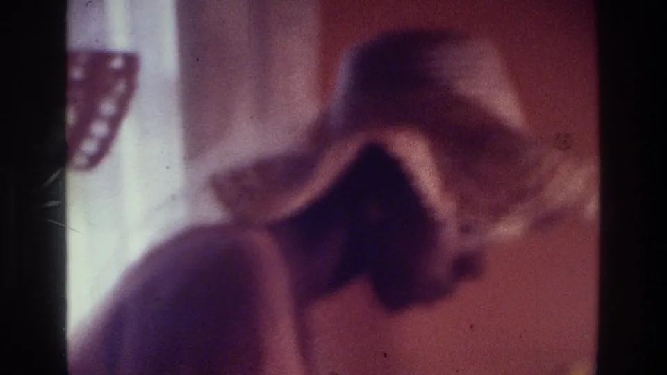 1975 Bronx home-movie frame of a woman in a straw hat leaning forward, with her face partly hidden by shadow.