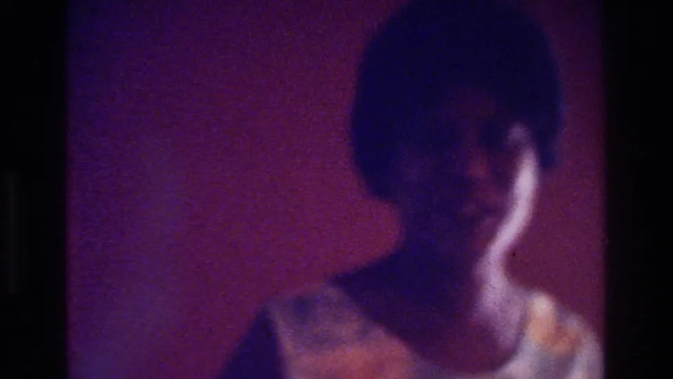 1975 Bronx home-movie still showing a dark-haired person in a light top, filmed in soft focus under reddish-purple indoor light.