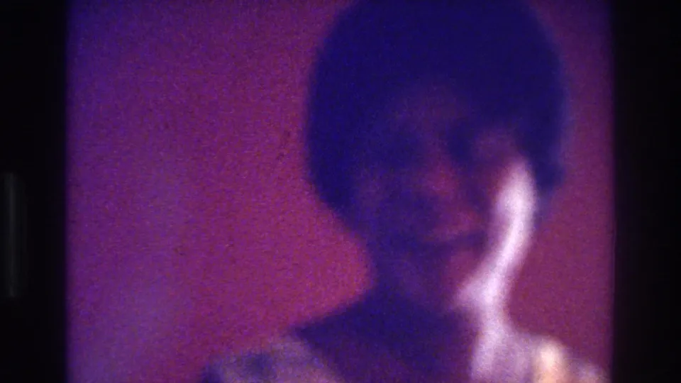 1975 Bronx 8mm still of a dark-haired person in a light top, seen close-up with strong purple color shift and grain.