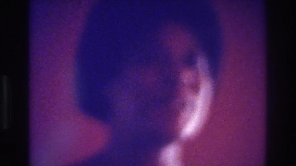 1975 Bronx home-movie frame with a soft profile-like view of a dark-haired person, purple-red background, and visible 8mm grain.