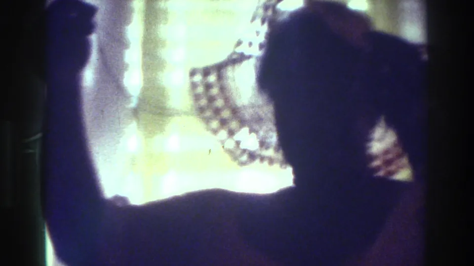 1975 Bronx home-movie frame showing a silhouetted person raising an arm near a bright window, with a patterned hat or head covering visible.