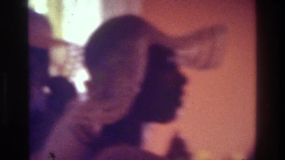 1975 Bronx home-movie frame with a woman in a patterned hat seen in silhouette beside warm wall light inside a private party room.