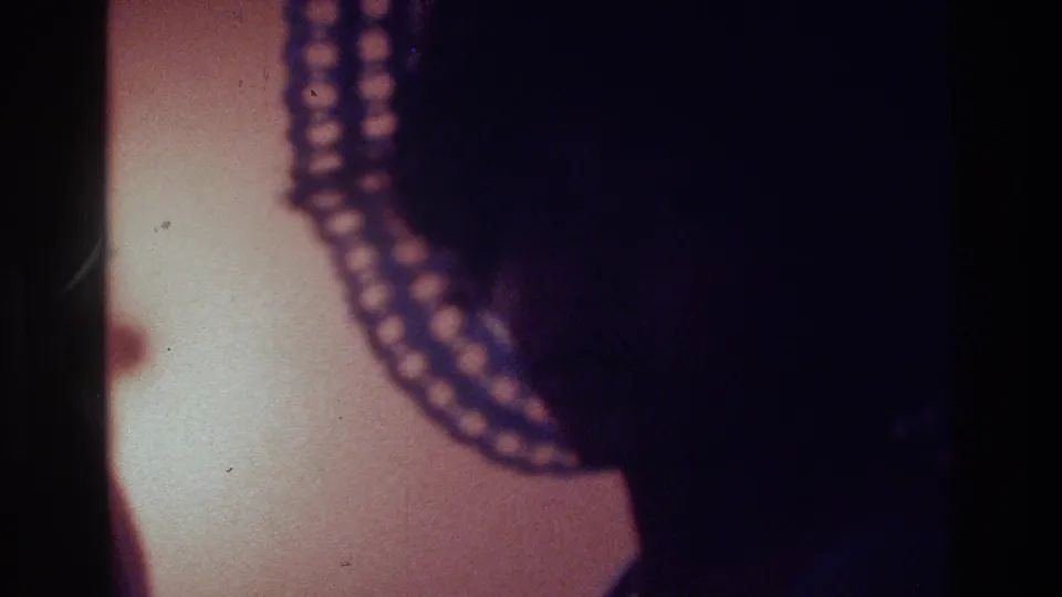 Close 1975 Bronx party frame showing layered beaded jewelry and a dark figure, with warm wall color and heavy 8mm grain.