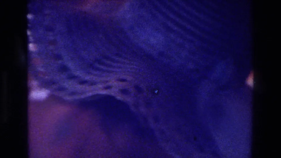Blue-shifted 1975 Bronx home-movie frame showing a close patterned hat or fabric detail in a dim private party interior.