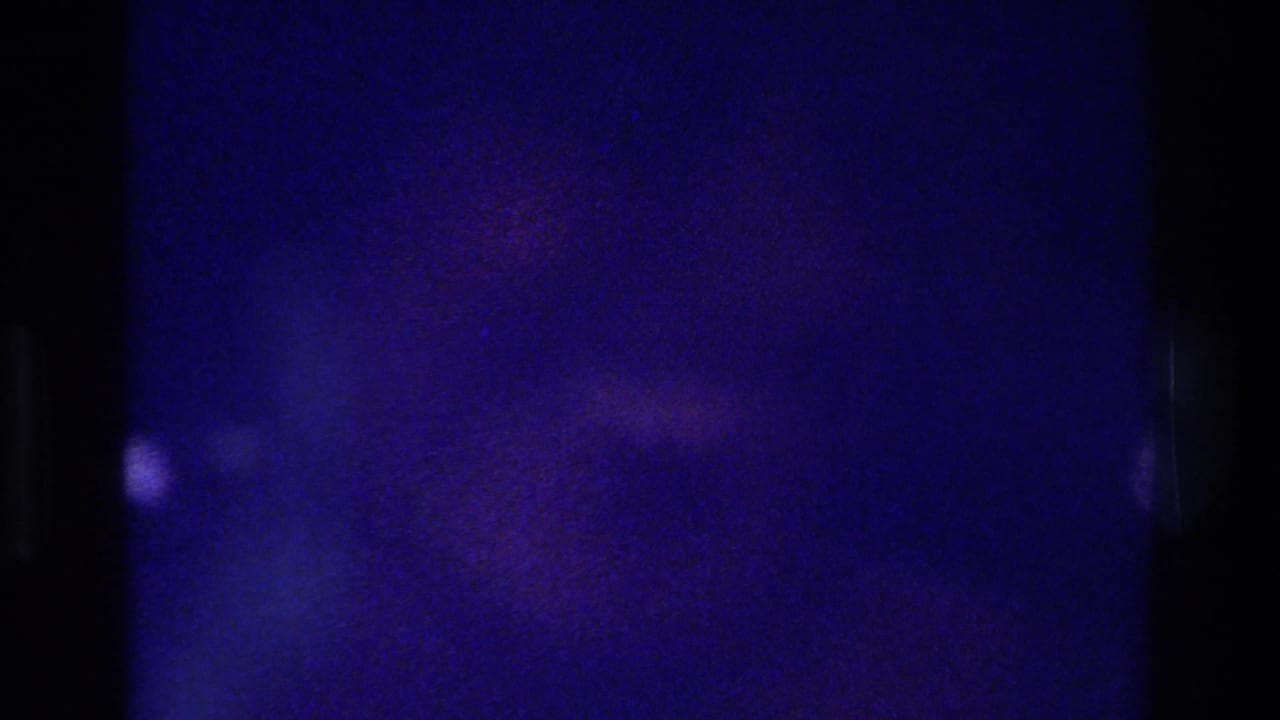 Dark blue-purple film frame with a small bright speck and heavy grain.