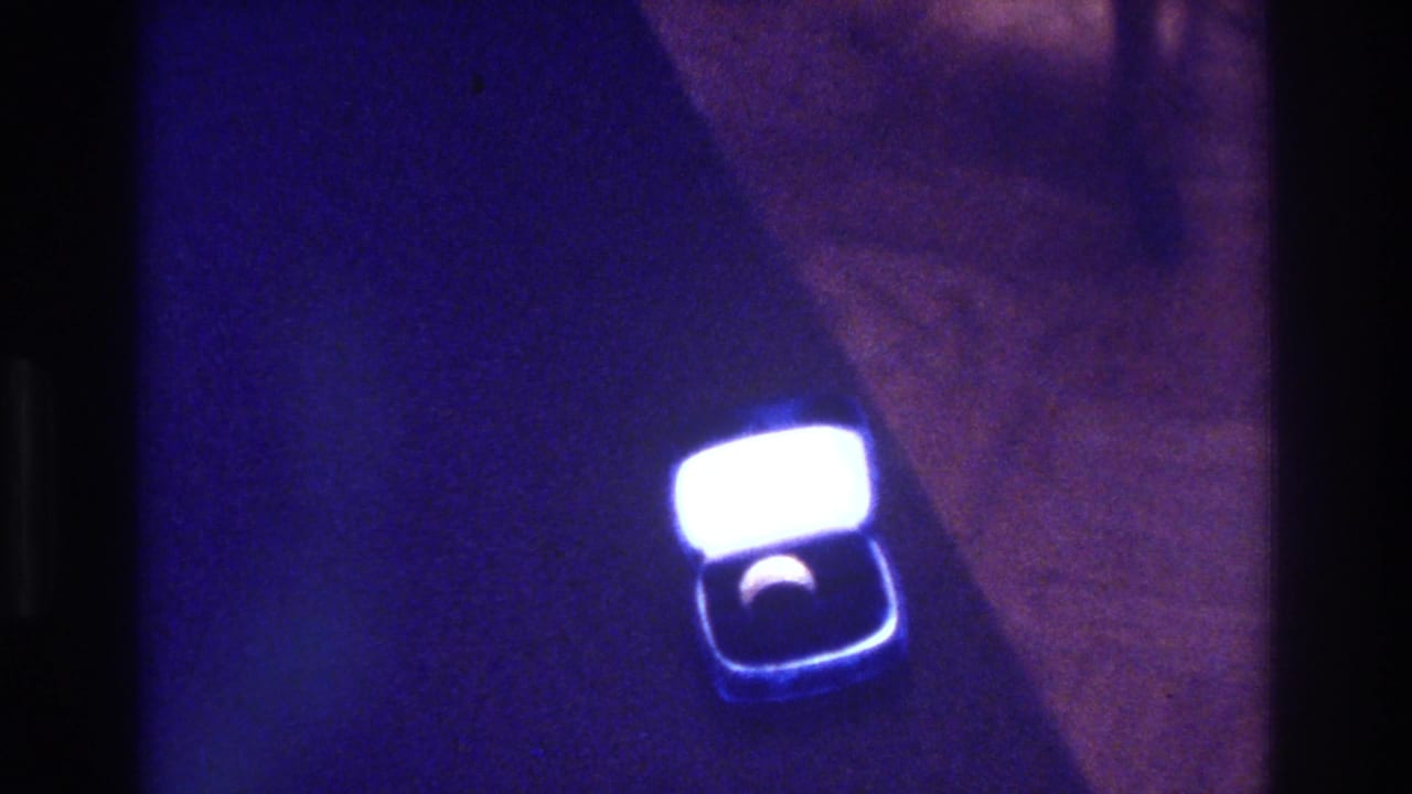 1975 Bronx 8mm home-movie frame showing an open square ring box with a bright glowing lid and a ring set into a dark interior under blue-purple light.