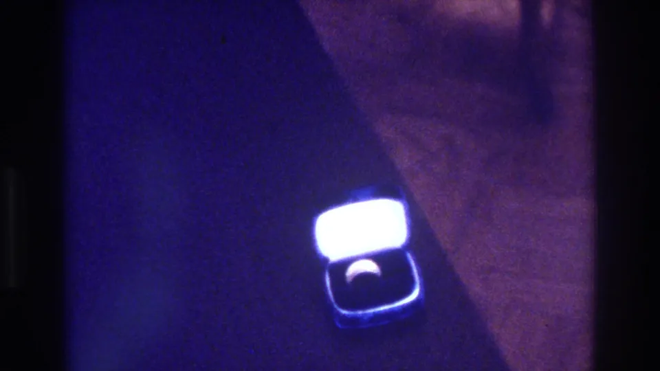 1975 Bronx 8mm home-movie frame showing an open square ring box with a bright glowing lid and a ring set into a dark interior under blue-purple light.