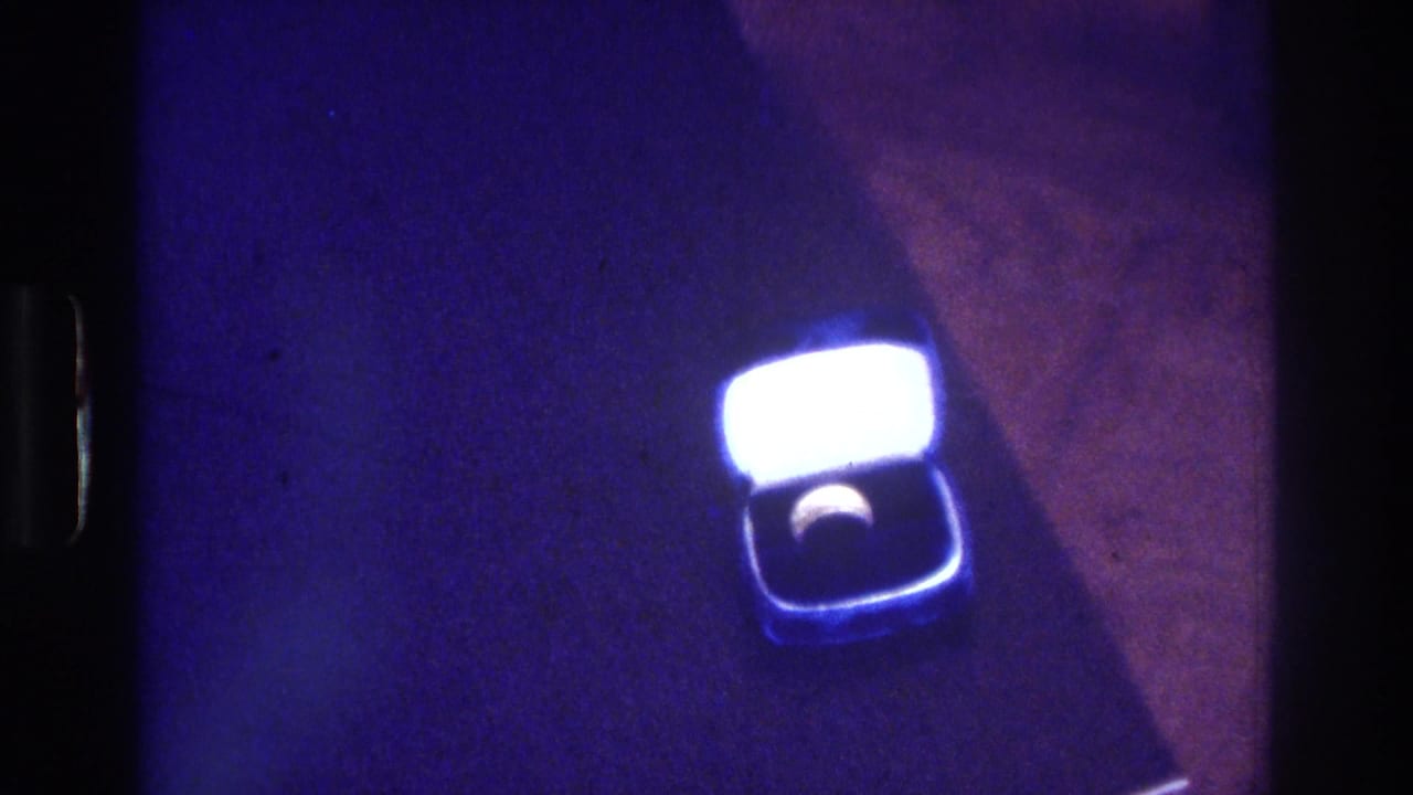 1975 Bronx 8mm frame with a small open ring box viewed from above, its lid glowing brightly while the ring sits in shadow inside the box.