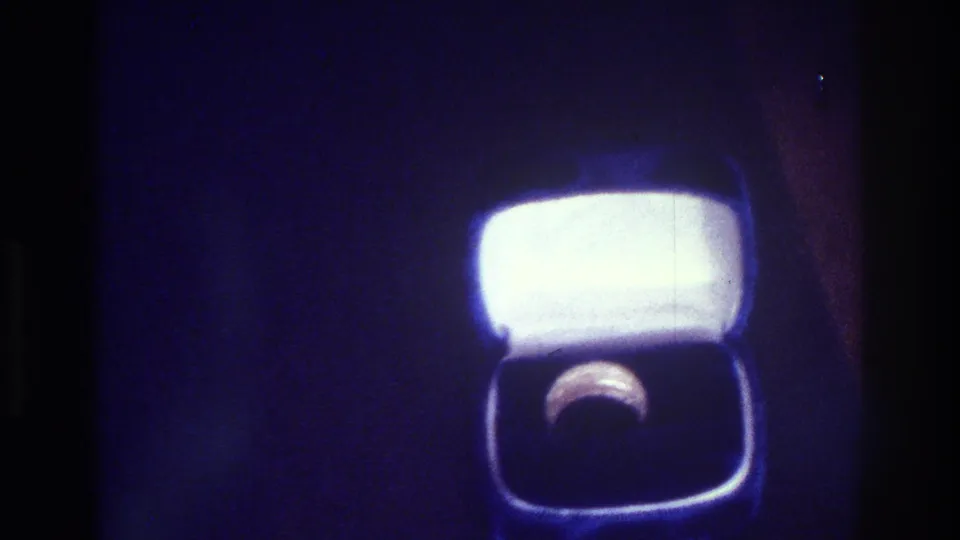 Closer 1975 Bronx 8mm home-movie view of a ring resting inside an open square box, with the bright lid blooming into the surrounding blue-purple exposure.