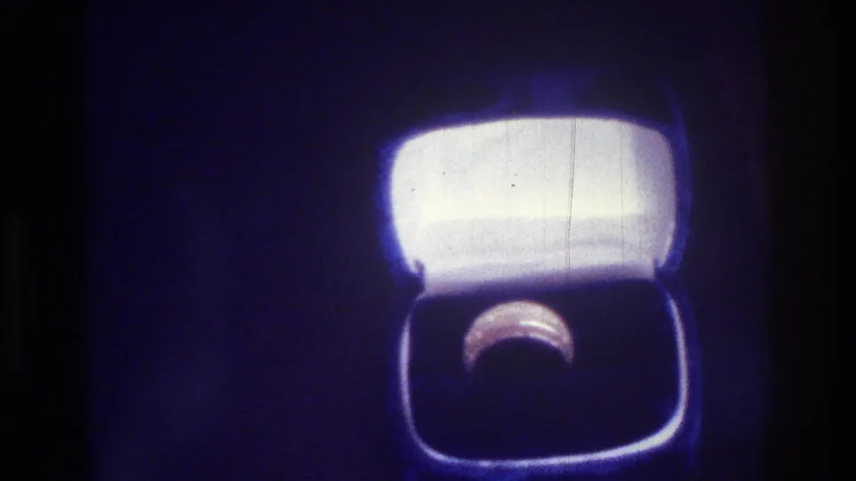 1975 Bronx 8mm close-up of an open wedding ring box, showing a bright white lid above a ring set into a dark lower compartment with soft blue-purple film color.