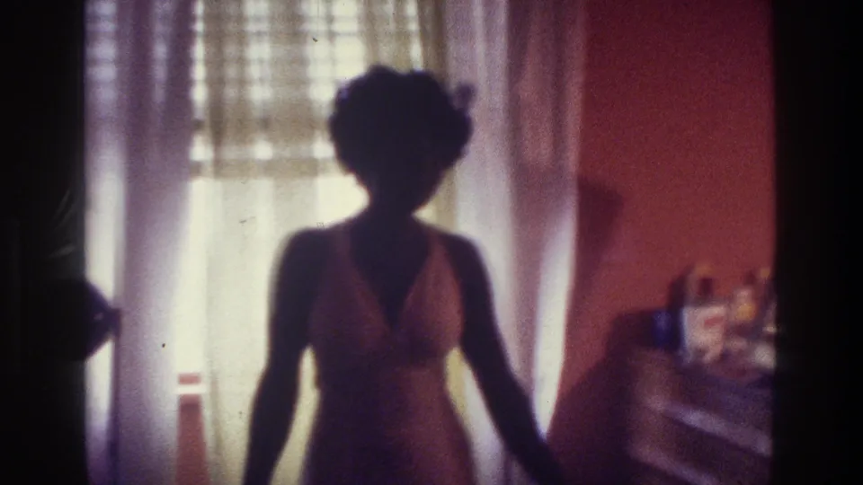 1975 Bronx 8mm frame showing a silhouetted woman in a halter dress inside a sunlit bedroom with sheer curtains.