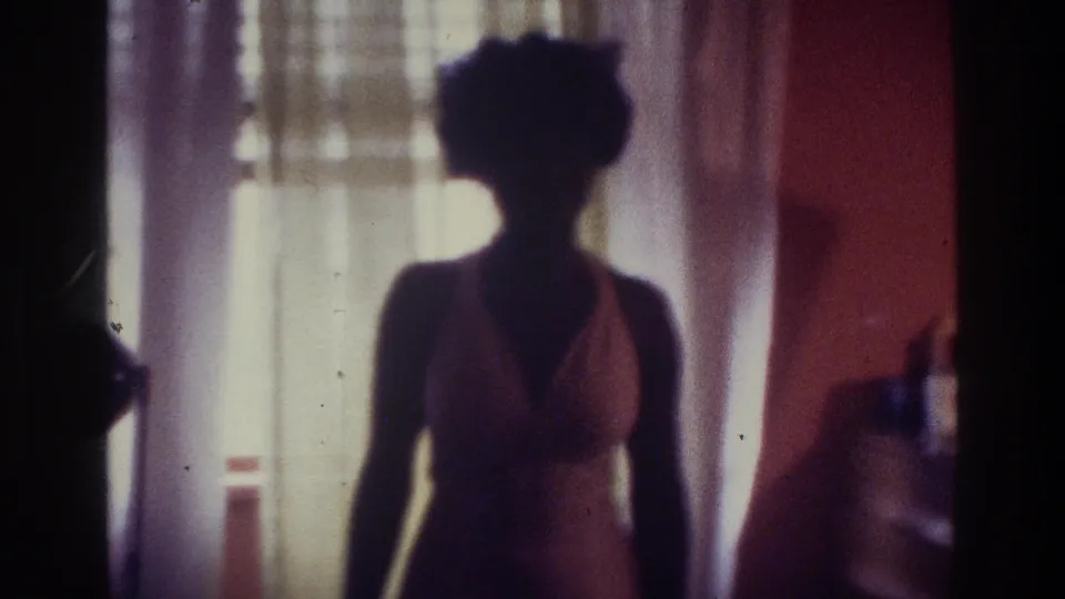 1975 Bronx 8mm frame of a woman silhouetted against sheer curtains in a warm private bedroom interior.