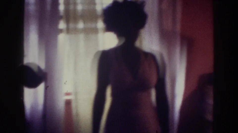 1975 Bronx home-movie frame showing a dark silhouette of a woman in a swimsuit near a bright curtained window.