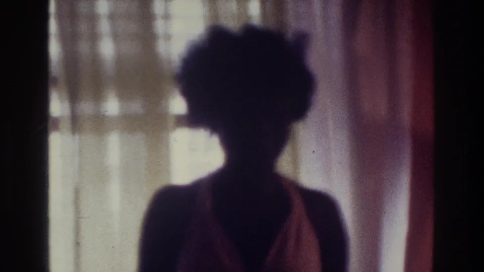 Soft-focus 1975 Bronx home-movie frame showing a closer swimsuit silhouette before bright indoor curtains.