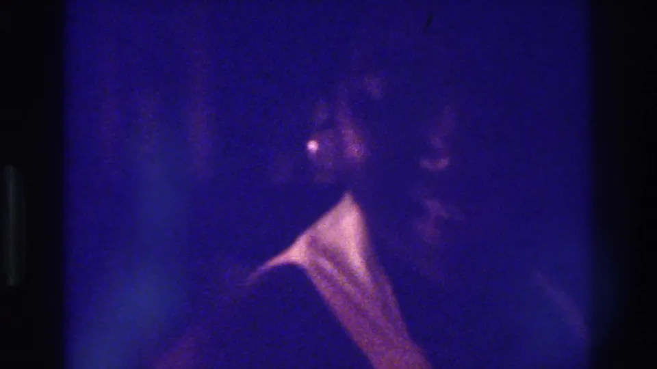 1975 Bronx 8mm frame showing a woman with a pale collar or neckline moving through a dark, grainy interior.