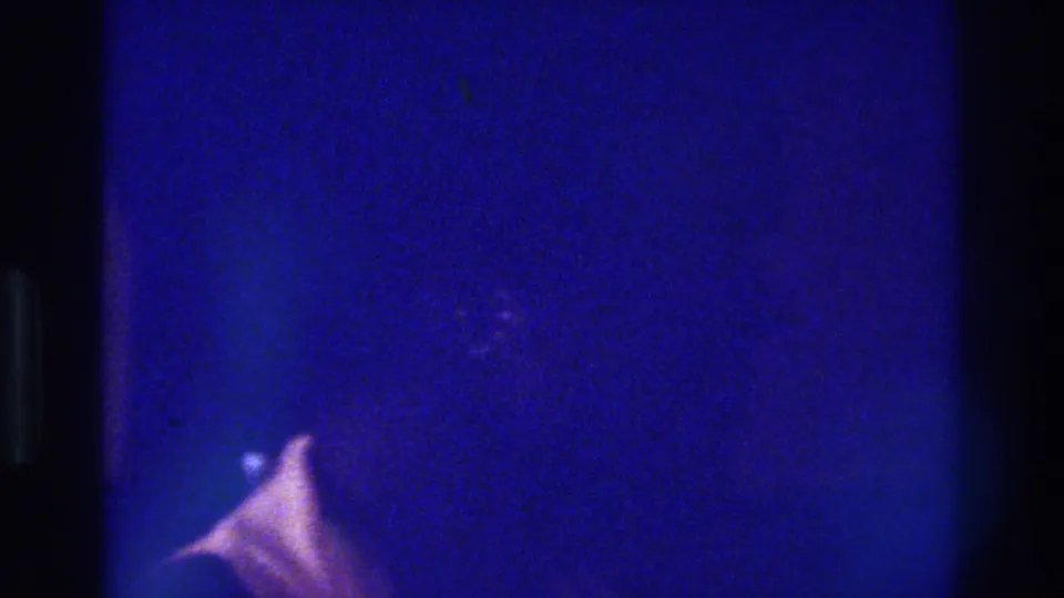 Low-light 1975 Bronx 8mm frame with a faint woman or figure receding into blue shadow and a small pale highlight below.
