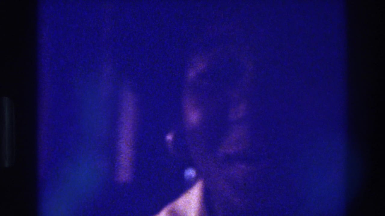 Low-light 1975 Bronx home-movie frame with an indistinct adult profile, blue cast, and pale garment highlight.