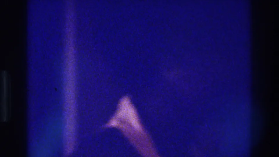 Final 1975 Bronx 8mm frame with blue-purple grain, a pale clothing shape, and a bright white flare near the top.