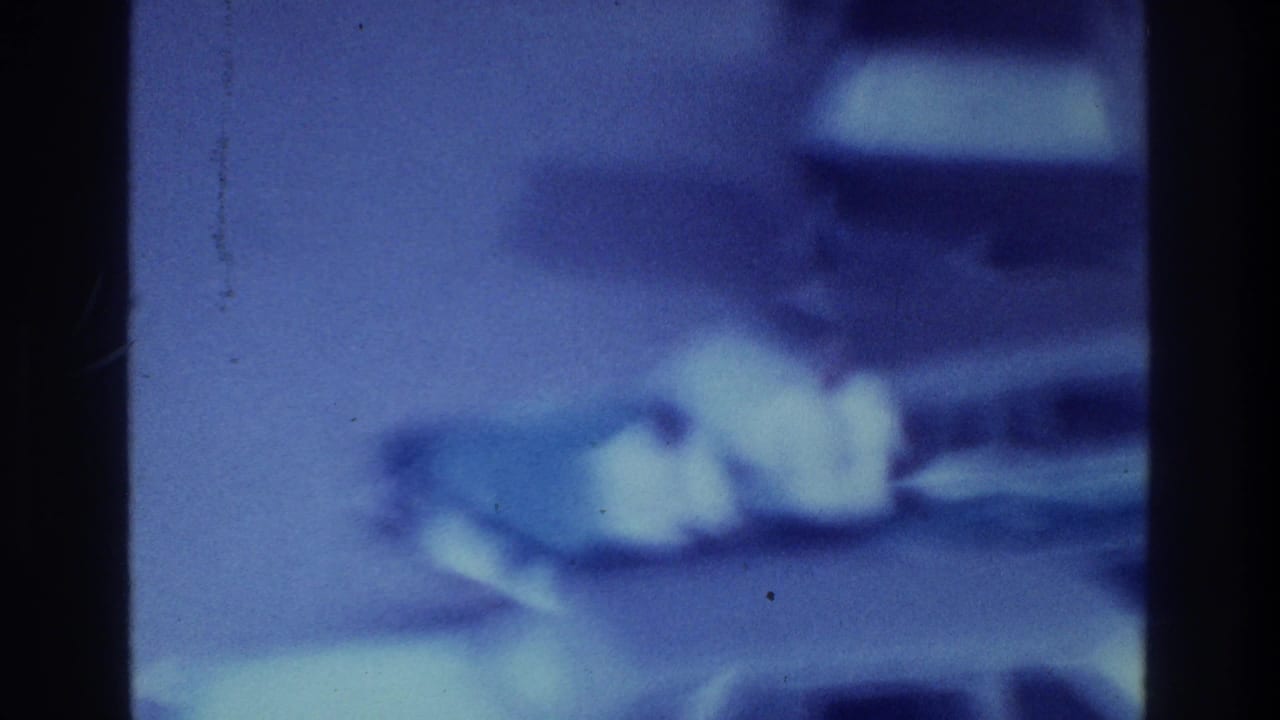 1975 Bronx home-movie frame showing a heavily blurred, blue-purple possible waterfront scene with pale indistinct shapes and film borders.