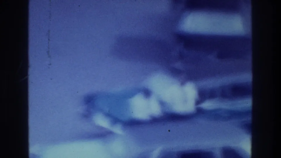 1975 Bronx home-movie frame showing a heavily blurred, blue-purple possible waterfront scene with pale indistinct shapes and film borders.