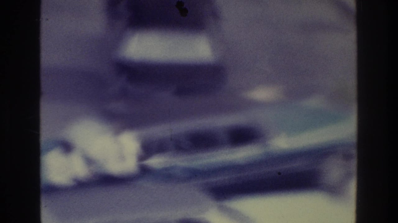 Soft 1975 Bronx home-movie frame showing a blurred vehicle on a road with heavy grain, gate edges, and blue-purple film fading.