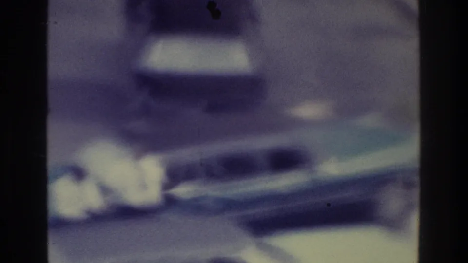 Soft 1975 Bronx home-movie frame showing a blurred vehicle on a road with heavy grain, gate edges, and blue-purple film fading.