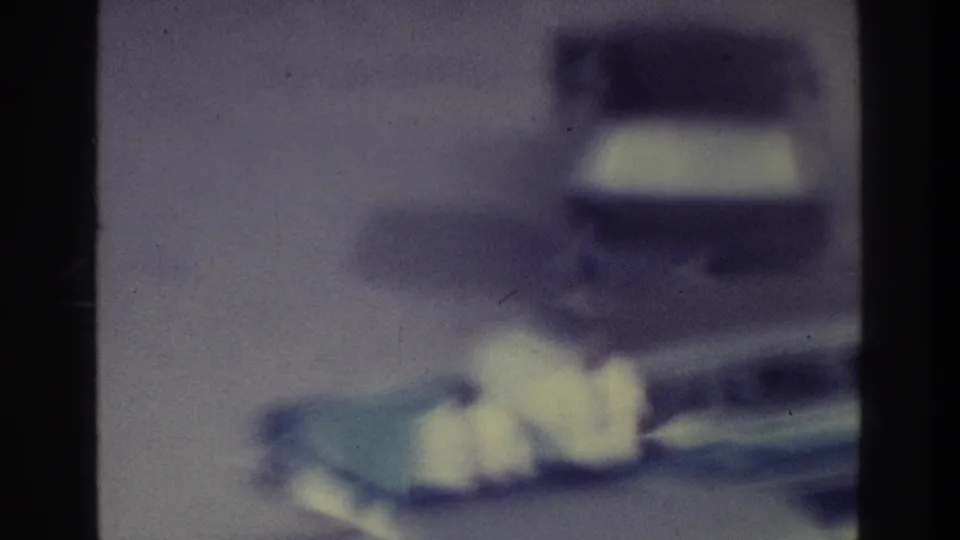 Blurred 1975 Bronx traffic frame with a boxy vehicle shape, roadway, pale foreground streaks, and aged 8mm color fading.