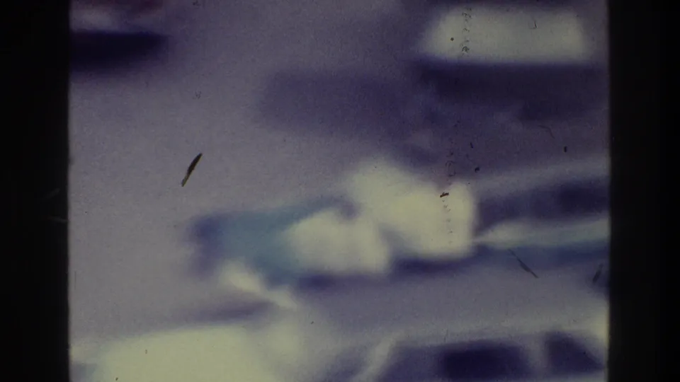 Soft-focus 1975 Bronx home-movie frame showing several parked cars, pavement, dust marks, and dark gate edges.