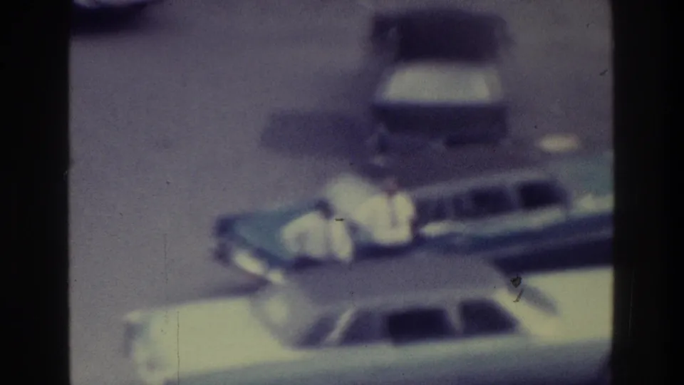 1975 Bronx 8mm frame with a light-colored sedan in the foreground and other parked cars behind, all softened by motion and grain.