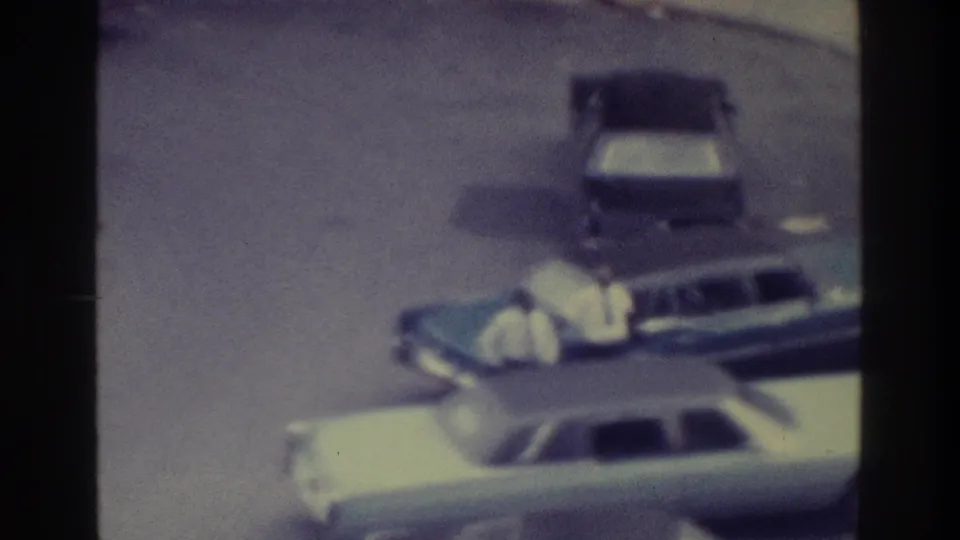 Color-shifted 1975 Bronx 8mm frame showing a light sedan, a darker parked car, open pavement, and visible film borders.