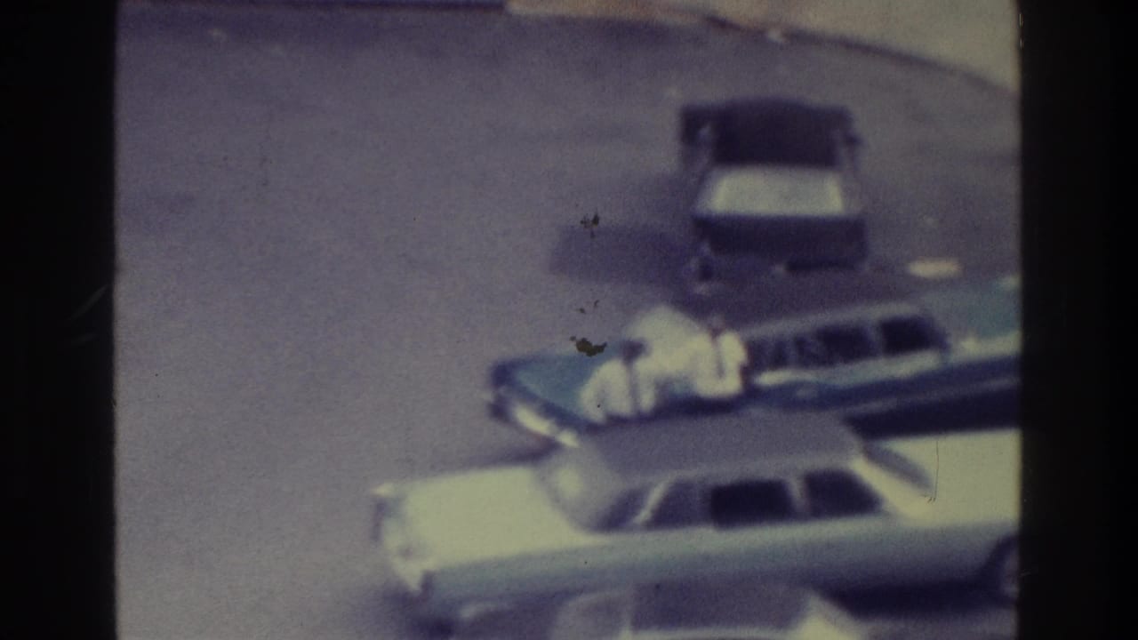 1975 Bronx 8mm frame showing a light sedan, dark station wagon, parked car, curb, and distant sidewalk pedestrians.