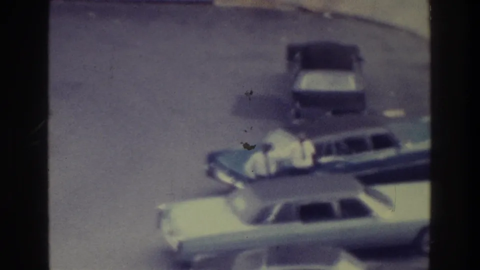 1975 Bronx 8mm frame showing a light sedan, dark station wagon, parked car, curb, and distant sidewalk pedestrians.