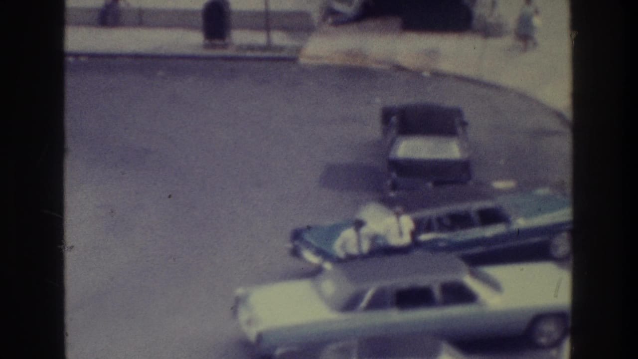 Parked light sedan and dark station wagon beside a curb, with pedestrians in the background.