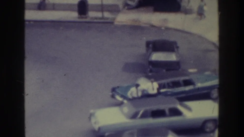High-angle 1975 Bronx home-movie frame with parked cars on asphalt, a person by the station wagon, and sidewalk figures beyond.