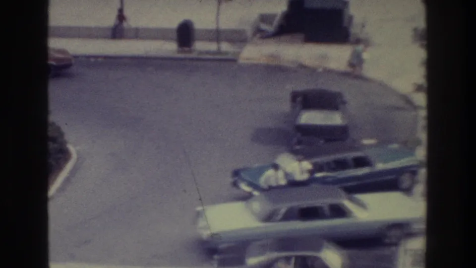 Faded 8mm Bronx frame from 1975 showing parked American cars, a curbside sidewalk, and small distant figures in an urban exterior.
