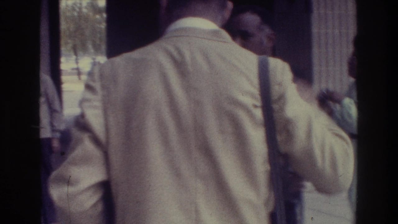Man in a light suit stands near a doorway as guests gather around him.