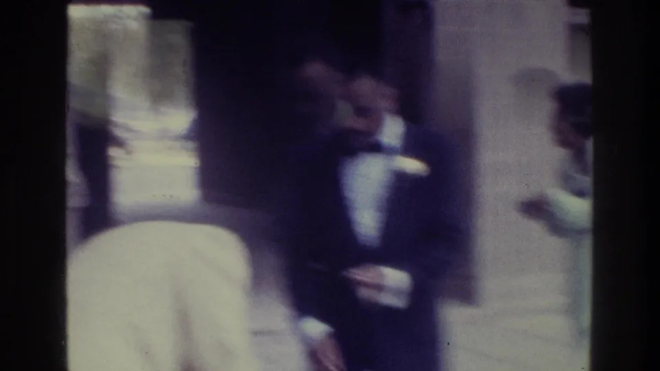 1975 Bronx wedding home-movie frame with guests in formal clothing gathered near a doorway and columned entrance.