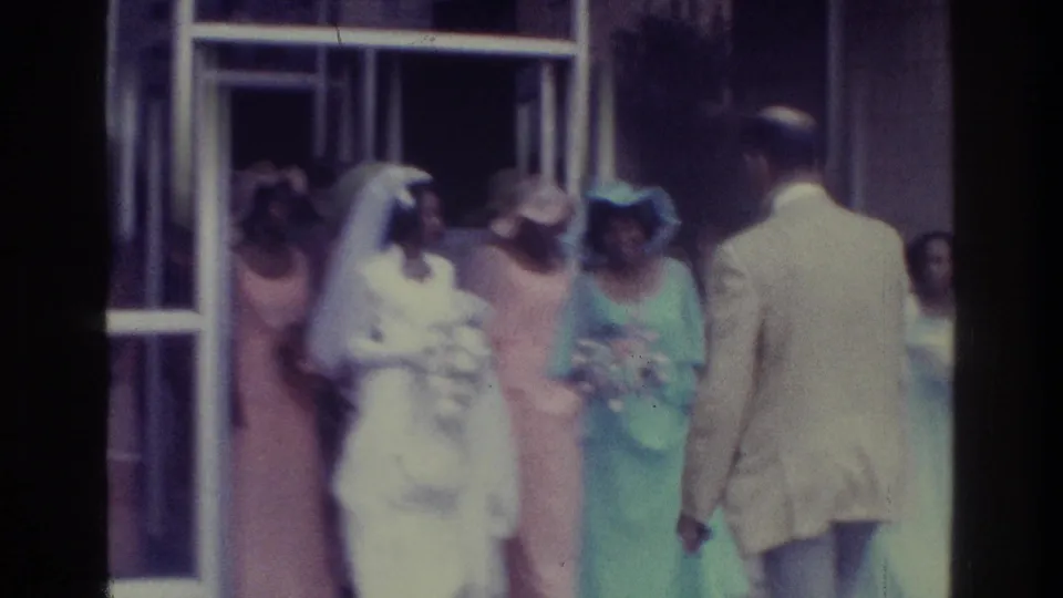 1975 Bronx wedding home-movie frame with bride, guests, open glass doors, and a man in a light suit leaving frame.