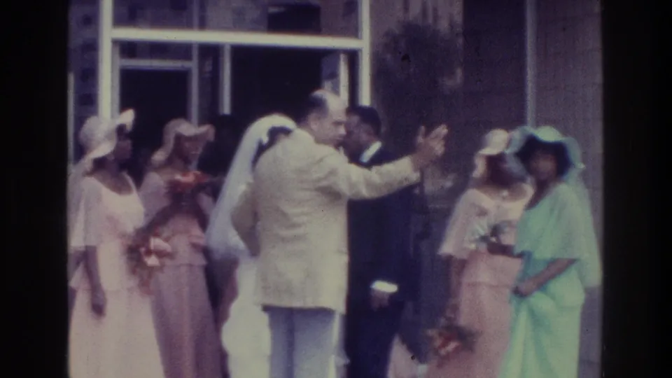 At a 1975 Bronx wedding exit, a man in a light suit greets a guest beside the bride and pastel-dressed attendants.