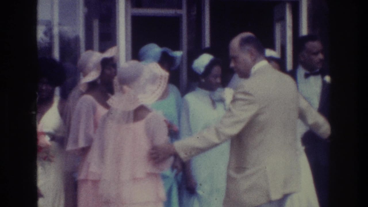 1975 Bronx 8mm frame showing a wedding party outside, with a man in a light suit arranging bridesmaids in pastel dresses and wide hats.