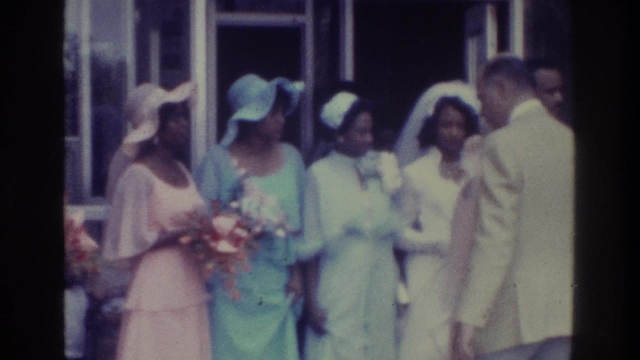 1975 Bronx wedding-party frame with bridesmaids in pastel dresses and hats standing while a man in a pale suit faces them.
