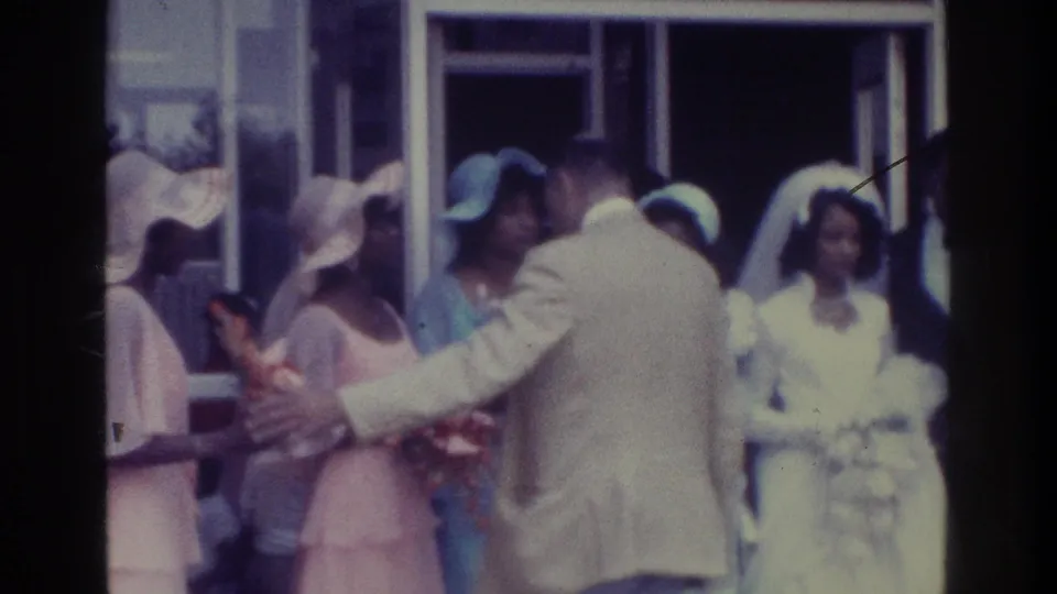 1975 Bronx 8mm frame showing a wedding-party arranger extending his arm as bridesmaids in pastel dresses shift positions.