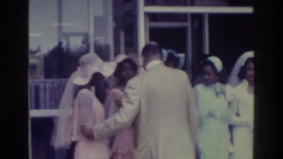1975 Bronx wedding-party frame with a man in a pale suit turned away while bridesmaids in pastel dresses face the camera area.